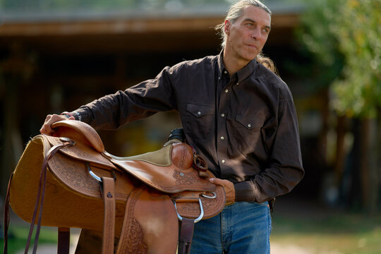 Man with long hair holding a brown leather saddle outdoors near a barn. Brandenburg, Germany