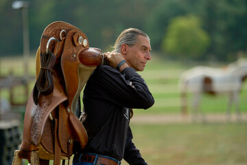 Man carrying a saddle over his shoulder in a pastoral setting with a horse in the background. Brandenburg, Germany