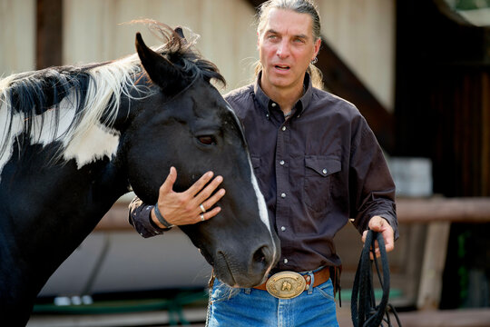 Man in denim and dark shirt affectionately holding a black and white horse outdoors. Brandenburg, Germany