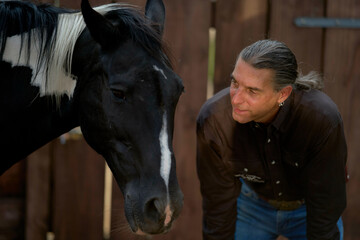 Man with long hair interacting with a black horse in a wooden stable setting. Brandenburg, Germany