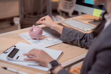 Businesswoman putting coin in piggy bank and using calculator to calculate savings and investments at workplace, managing home budget and analyzing financial data