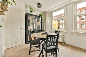 A stylish dining area featuring a round table, black chairs, and a bright, airy atmosphere with natural light streaming through large windows.