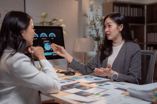 Two asian businesswomen are discussing company performance and analyzing financial charts displayed on a computer screen and printed documents, working together in a modern office