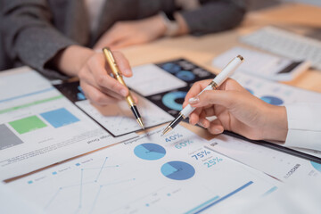 Two businesswomen are collaborating on analyzing financial charts and graphs, using pens to point...