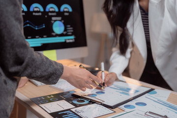Two businesswomen analyzing financial charts and graphs on a computer screen and printed documents while discussing company strategy and future investments in a collaborative office