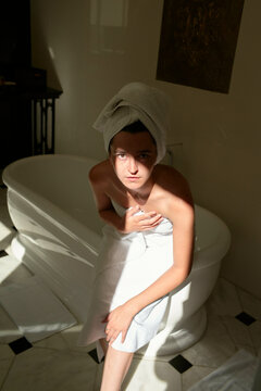Woman in towel sitting on bathtub in sunlit bathroom with marble floor.