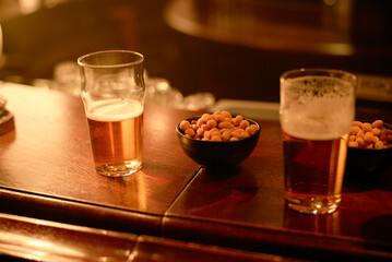 Two glasses of beer and bowls of peanuts on a wooden bar counter in a dimly lit pub, Vilnius, Lithuania