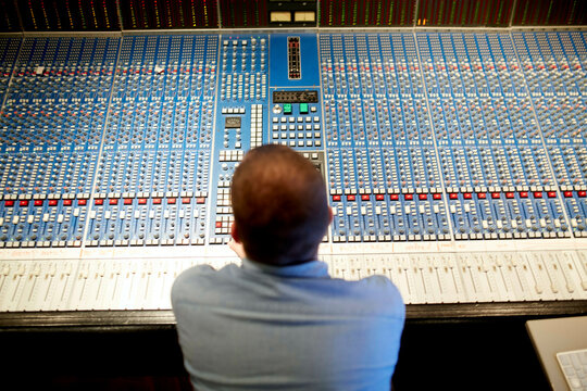Man in blue shirt operating a large audio mixing console in a recording studio.