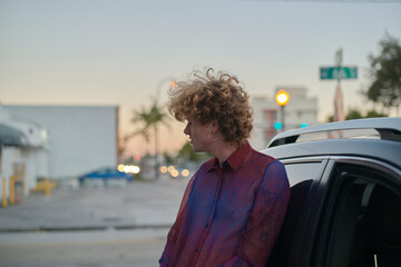 Person with curly hair leans against car door on an urban street at twilight. Miami, FL, USA