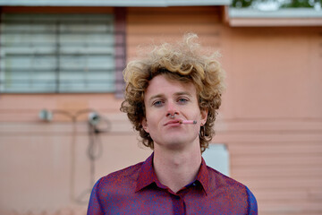 Young man with curly hair wearing a red shirt and smoking outside a peach-colored building. Miami, FL, USA