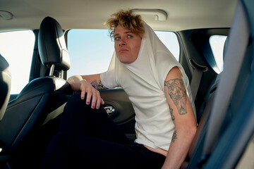 A person with tattoos sits in a car backseat, wearing a white shirt pulled over their head. Miami, FL, USA