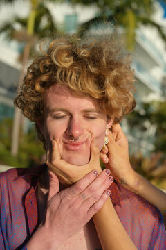 Young man with curly hair smiles as hands playfully squish his cheeks outdoors. Miami, FL, USA