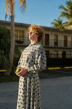 Person in a patterned dress holding an avocado stands outside a building with palm trees. Miami, FL, USA