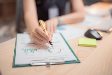 Businesswoman is analyzing financial charts and graphs using pen, sitting at her desk in the office, with a computer mouse and a sticky note nearby