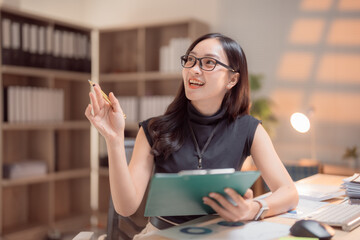 Asian businesswoman sitting at her desk, holding a clipboard and explaining her strategy during a...