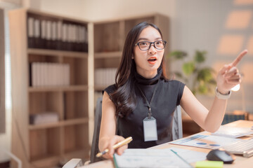 Asian businesswoman wearing glasses and lanyard with blank badge is holding pen and pointing finger...