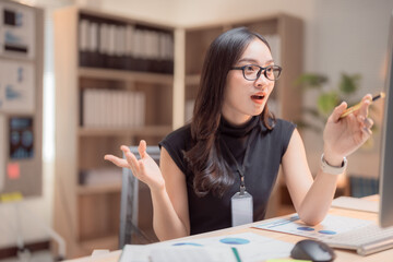 Young businesswoman wearing glasses sitting at her desk explaining graphics showing on her computer screen during a video call, holding a pen and gesturing with her hands