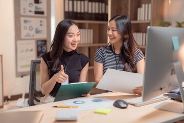 Two happy Asian businesswomen are analyzing financial charts and company sales documents, using a computer and showing thumbs up for good news