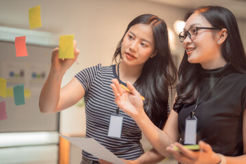 Two young businesswomen collaborating on a project, using sticky notes on a glass wall during a brainstorming session, generating innovative ideas and strategies together