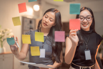 Two asian businesswomen are using colorful sticky notes attached to a glass wall while...