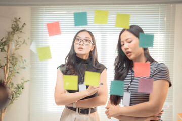 Two asian businesswomen are using colorful sticky notes on a glass wall during a brainstorming session in a modern office, discussing and sharing ideas for their next project