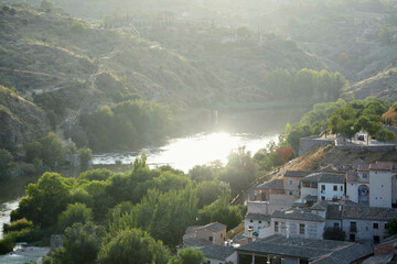 Scenic view of a sunlit river with trees and hillside village buildings in the foreground. Toledo, Spain