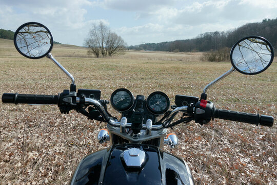 Motorcycle handlebars with mirrors reflecting a scenic view of a field and trees.