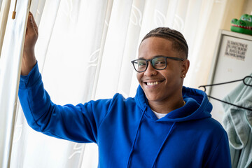Smiling young man in glasses and blue hoodie standing by window with sheer white curtains. Arnhem, Netherlands