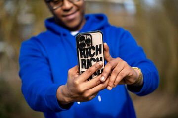 Man in blue hoodie taking a selfie with phone case reading RICH outdoors Arnhem, Netherlands