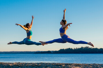 Two women in athletic attire perform graceful leaps on a beach at sunrise. Bainbridge Island, WA, USA