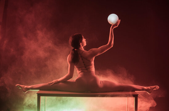 Dancer in a split on a table holds a glowing ball amidst colorful smoke.