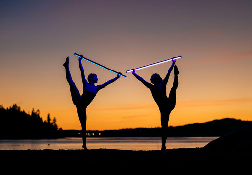 Two dancers perform with light sabers by a lake at sunset, silhouetted against colorful sky. Bainbridge Island, WA, USA