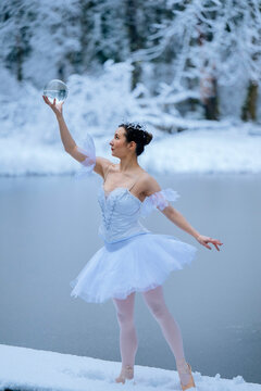 Ballerina in white tutu and tiara poses with a glass orb by a snow-covered lake. WA, USA