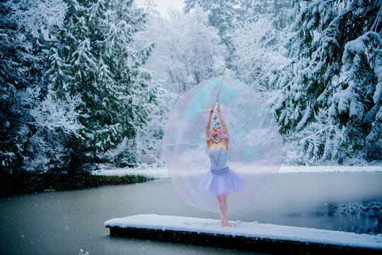 Ballerina in tutu posing on snow-covered dock with a large iridescent umbrella by a snowy lake. WA, USA