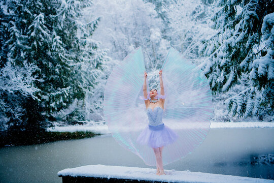 Woman in fairy costume stands on snowy dock by frozen lake surrounded by snow-covered trees. WA, USA