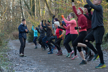 A group of diverse people dressed in athletic clothing exercises energetically on a wooded trail, guided by an instructor. Sale Water Park, Manchester, UK