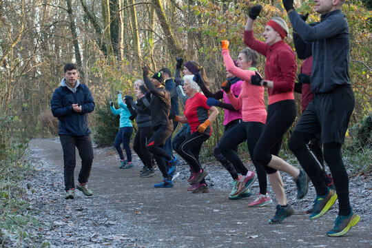 A group of people dressed in athletic gear are jogging on a forest path, led by a male instructor. Sale Water Park, Manchester, UK