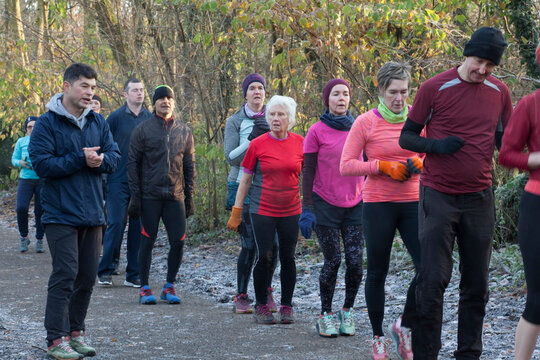 A diverse group of adults dressed in athletic wear participate in an outdoor group exercise in a forest setting. Sale Water Park, Manchester, UK