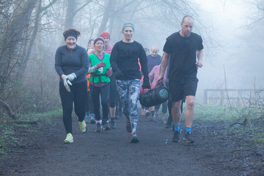 A group of diverse adults jogs on a forest trail in misty weather, dressed in colorful activewear. They are visibly energetic and focused. Sale Water Park, Manchester, UK
