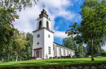 Fototapeta premium The Church of Sweden stands tall against a blue sky and clouds in Idre Dalarna. The little white wooden church and cemetery framed by lush green trees and vibrant fresh lawn