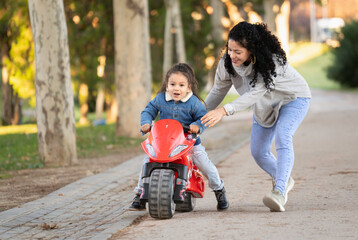 Latin mother helping her son riding toy motorcycle in park