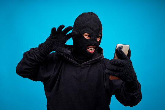 Cruel Man In Black Clothes With Hidden Face Looks At Smartphone Screen Isolated On Blue Background