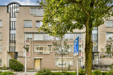 A contemporary residential building featuring large windows and a green exterior. The surrounding area includes trees and a parking sign, enhancing the urban environment.