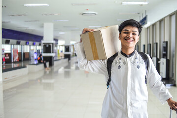 Happy Young Muslim Man Carrying Cardboard Box on Shoulder at Terminal, Ready To Go For Mudik