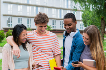 Four diverse young friends joyfully sharing a moment together while looking at their smartphones and laughing in a vibrant outdoor setting during a sunny day of their university life