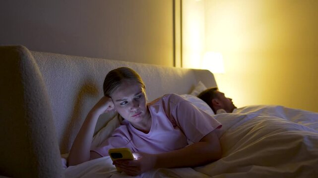Woman secretly texting on smartphone while husband sleeps in bed, infidelity, betrayal in marriage. Relationship issues, wife immersed in phone after argument, emotional distance, trust broken