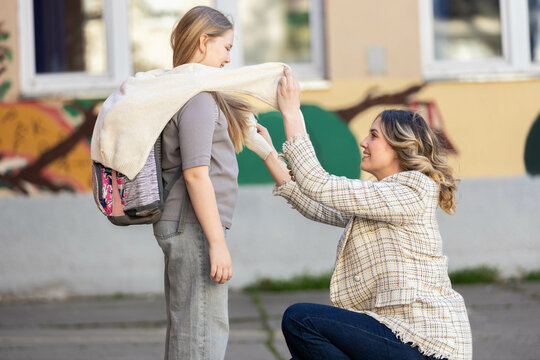 A mother gently assists her daughter by draping a sweater over her shoulders during a warm day at school.