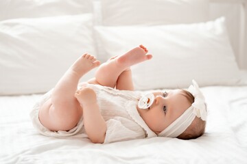 Cute newborn baby girl sucks a pacifier in white clothes on the bed at home on her back, cute baby...