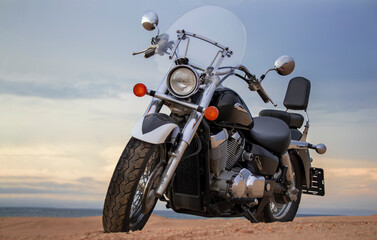 A lone motorcycle stands on the sandy surface of the Namib Desert against a dramatic cloudy sky. The sunset lighting highlights the contrast between nature and machine.