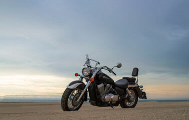A lone motorcycle stands on the sandy surface of the Namib Desert against a dramatic cloudy sky. The sunset lighting highlights the contrast between nature and machine.